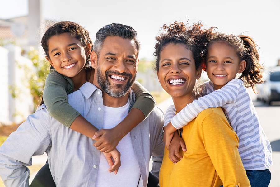 A smiling family of four outdoors, both parents giving a daughter a piggyback ride